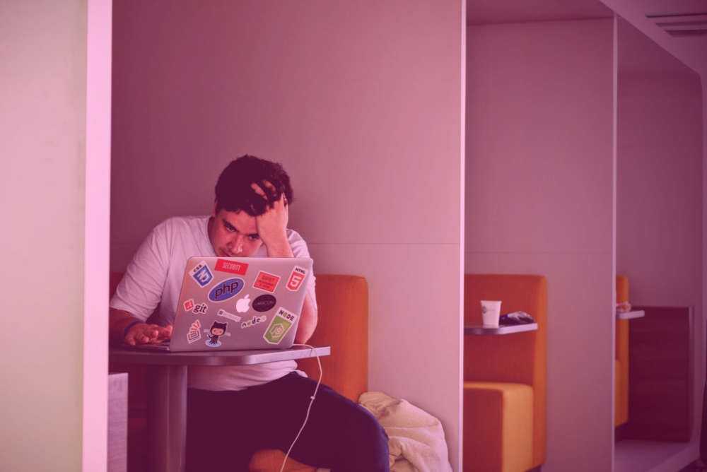 Man sitting in an orange booth at a silver table, fingers in his hair with a look of frustration, using his laptop