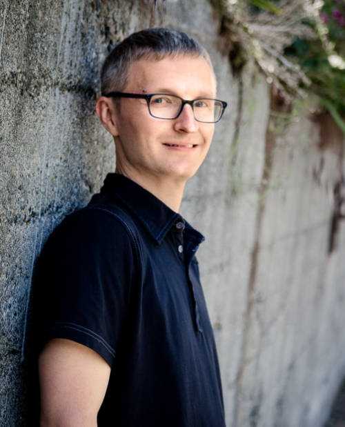 Devon wearing a black shirt and black glasses, standing against a gray retaining wall, smiling at the camera